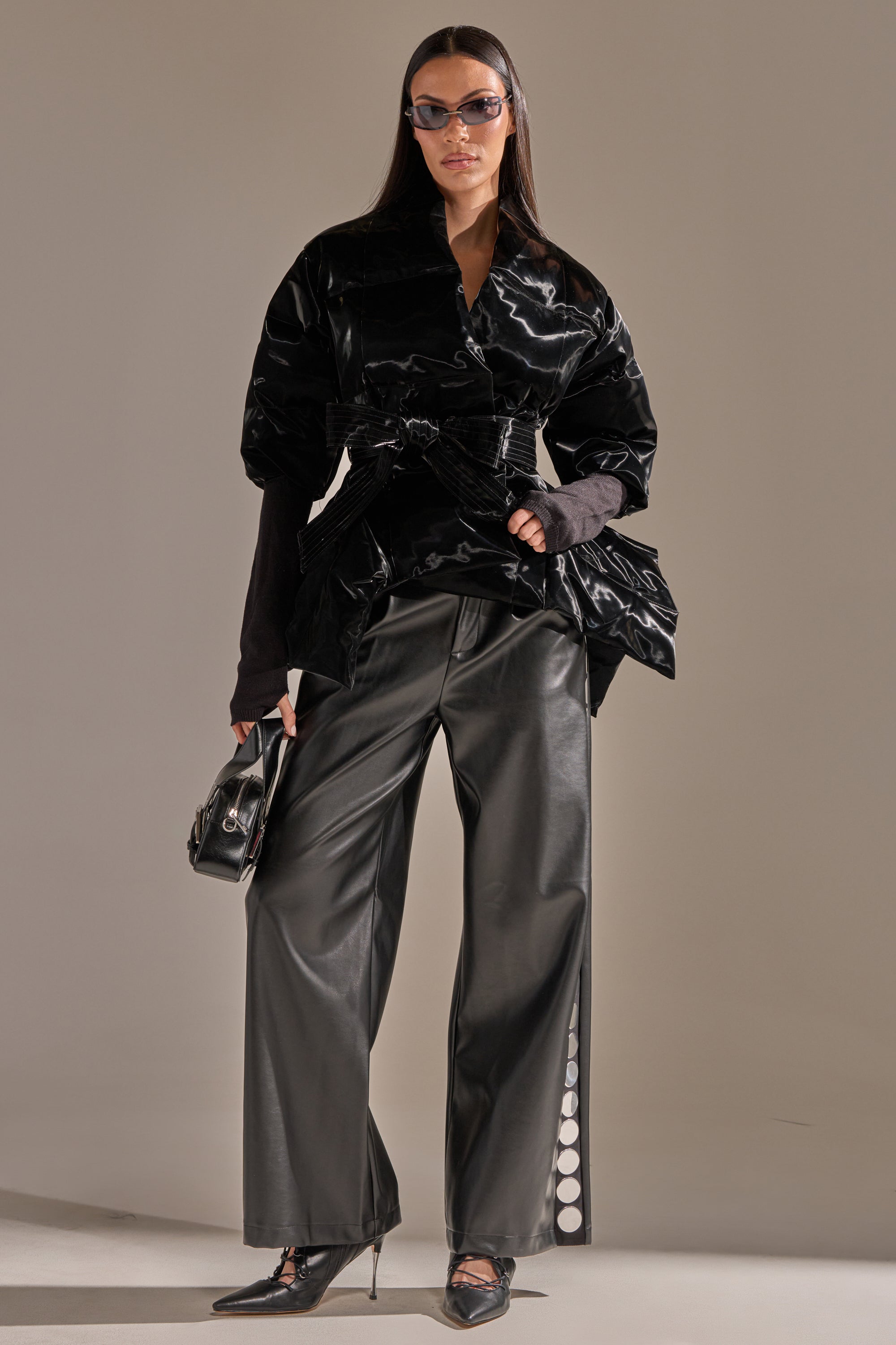 A woman stands confidently against a neutral background, wearing black sunglasses, pointed heels, wide-leg black leather pants, holding a small handbag—and the RIB SLEEVE PEPLUM PUFFER IN LIQUID VELVET. 7