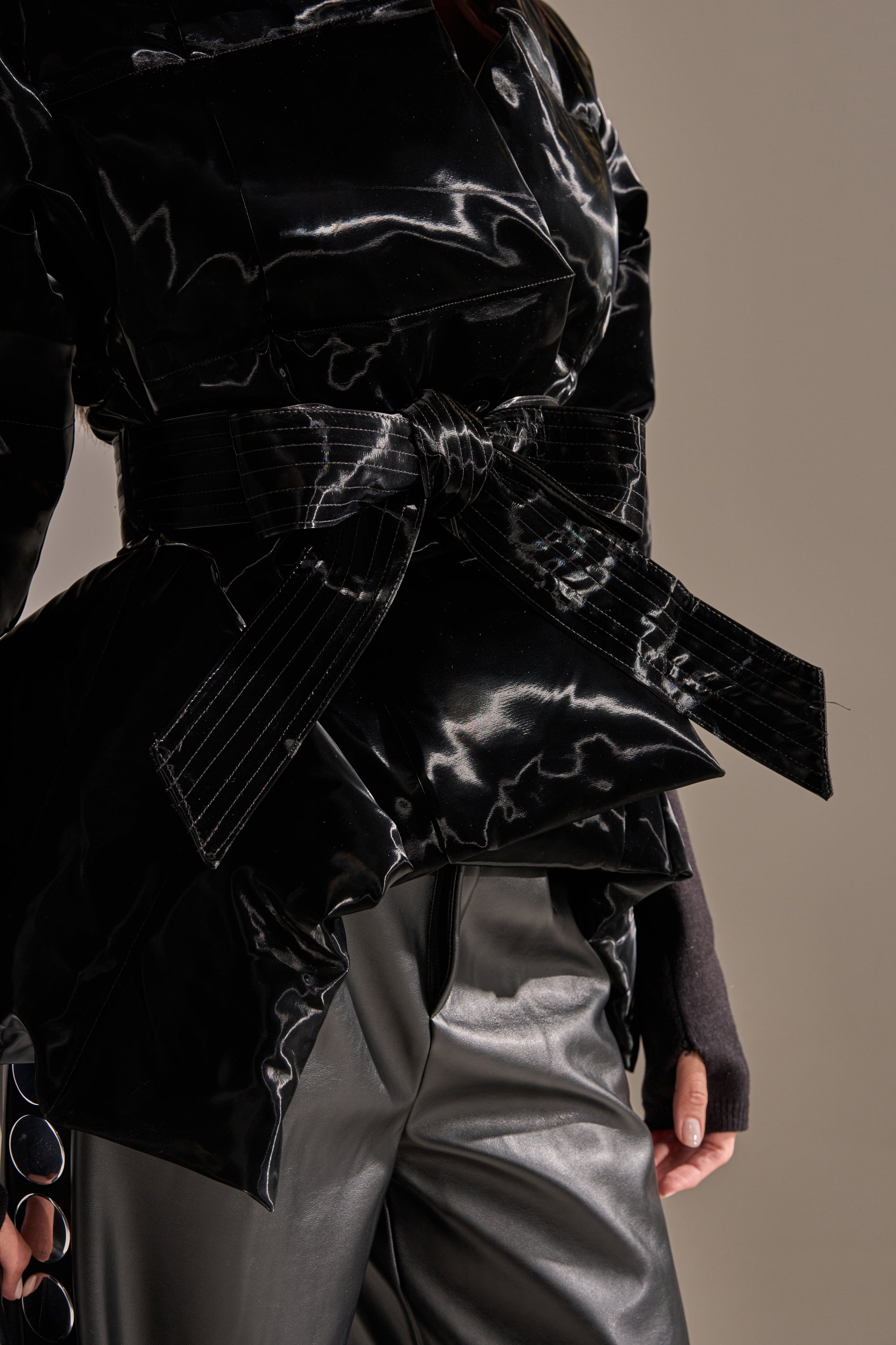 Wearing the RIB SLEEVE PEPLUM PUFFER IN LIQUID VELVET in glossy black, featuring a wide belted waist and paired with matching black pants, this look is set against a plain background with the right hand partly visible. 8
