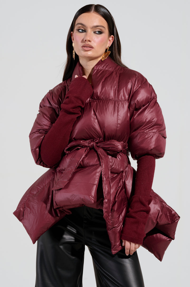 A person wearing the RIB SLEEVE PEPLUM PUFFER IN BURGUNDY, featuring a belt tied at the waist and long ribbed sleeves, pairs it stylishly with black leather pants while posing confidently against a plain background.