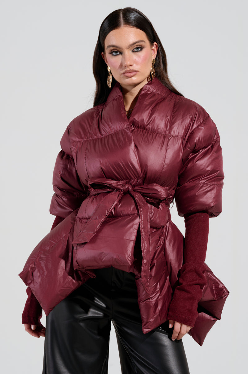 A woman with long dark hair confidently poses against a plain background, wearing the stylish RIB SLEEVE PEPLUM PUFFER IN BURGUNDY, complete with a tied waist. She pairs the chic jacket with black leather pants.