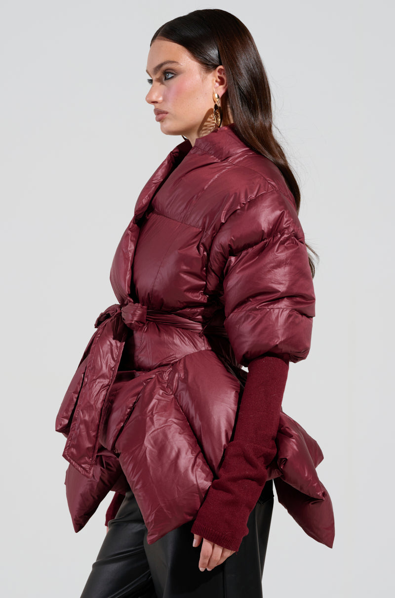 A woman with long, dark brown hair is looking to the side while wearing the RIB SLEEVE PEPLUM PUFFER IN BURGUNDY, which features an asymmetrical design and belt. She pairs the jacket with ribbed sleeves and black pants, all set against a plain white background.