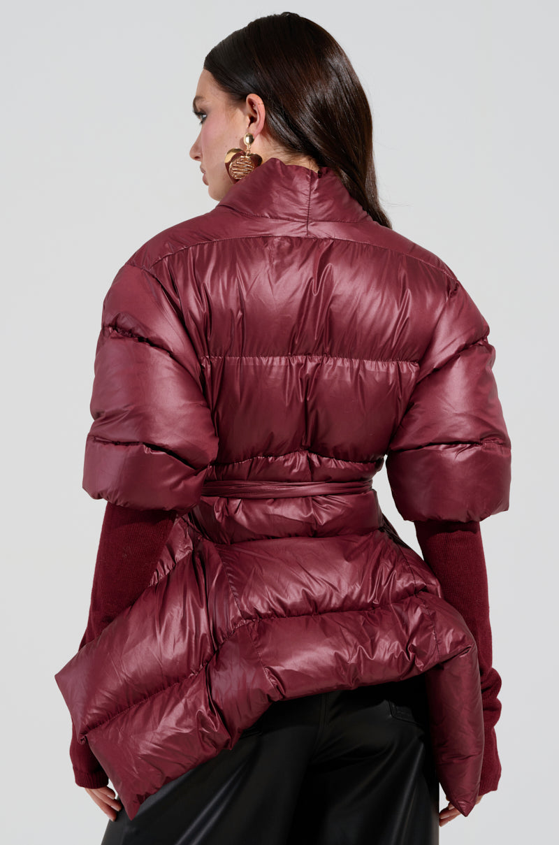 A person with long dark hair is seen from behind, wearing the RIB SLEEVE PEPLUM PUFFER IN BURGUNDY and a matching dark red sweater. The jacket features ribbed sleeves, a high collar, and is cinched at the waist. The plain light gray background enhances the sleek silhouette.