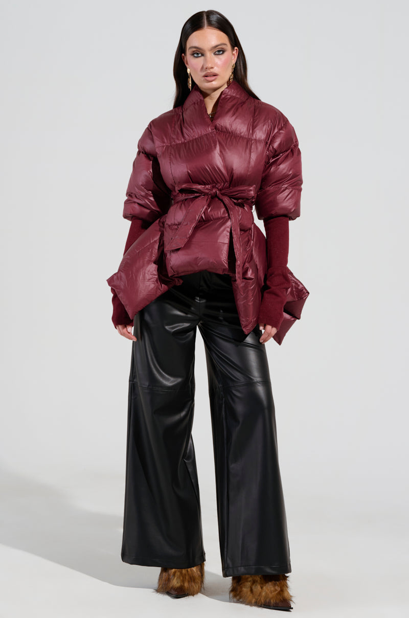 A person wearing the RIB SLEEVE PEPLUM PUFFER IN BURGUNDY with ribbed sleeves poses against a plain background, paired with wide-leg black leather pants featuring fur trim at the hem. The jacket has a belted waist, and the individual, sporting long dark hair, stands confidently.