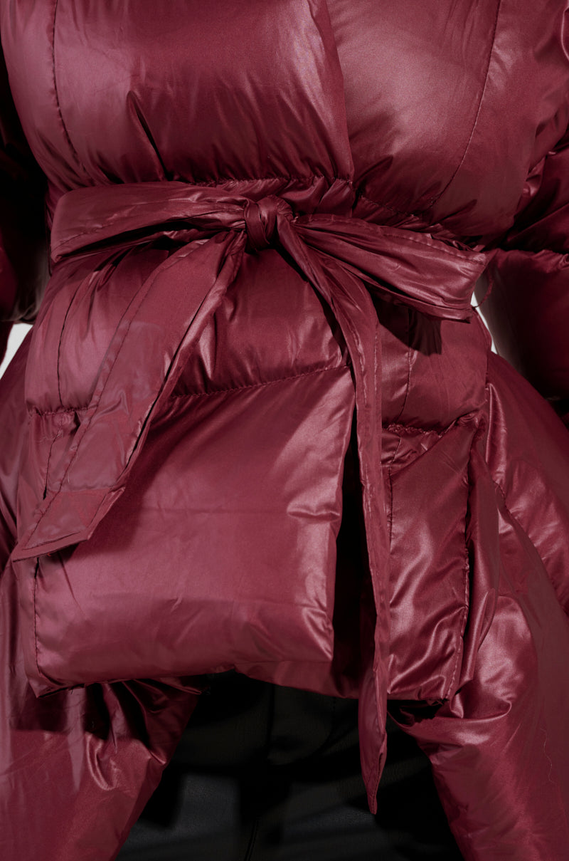 Close-up of the RIB SLEEVE PEPLUM PUFFER IN BURGUNDY, featuring a tied belt at the front, quilted texture, shiny fabric, and elegant ribbed sleeves.