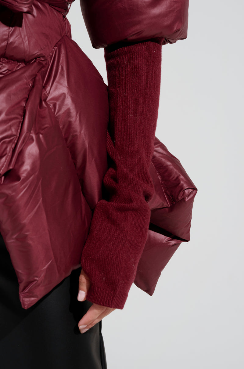 A close-up shows an individual wearing the Rib Sleeve Peplum Puffer in Burgundy, complemented by matching burgundy knit arm warmers. The ensemble highlights rich fabric textures, set against a neutral background.