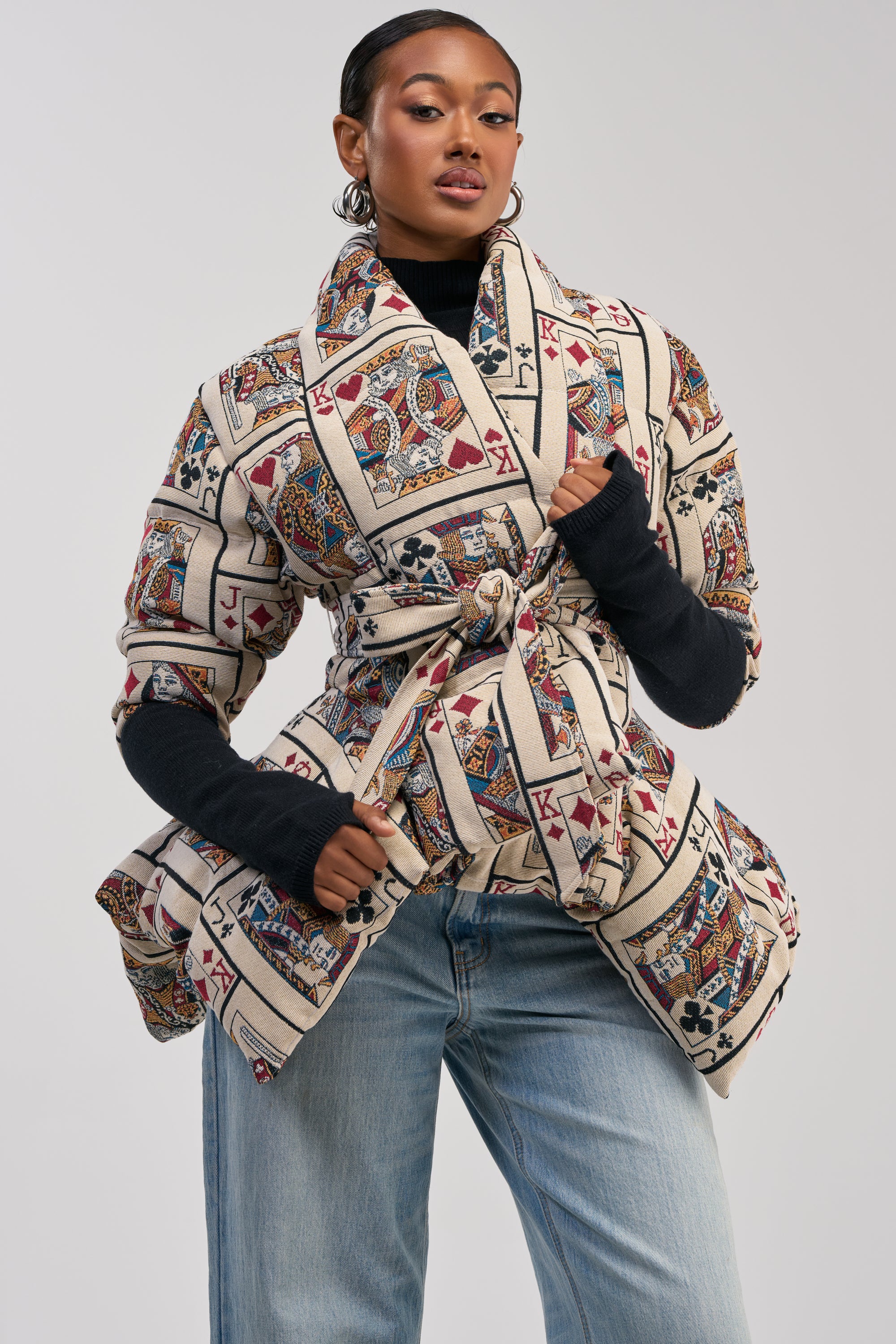 A woman poses confidently wearing the SHOW YOUR CARDS TAPESTRY PEPLUM PUFFER jacket layered over a black turtleneck and paired with light blue jeans, against a plain background.