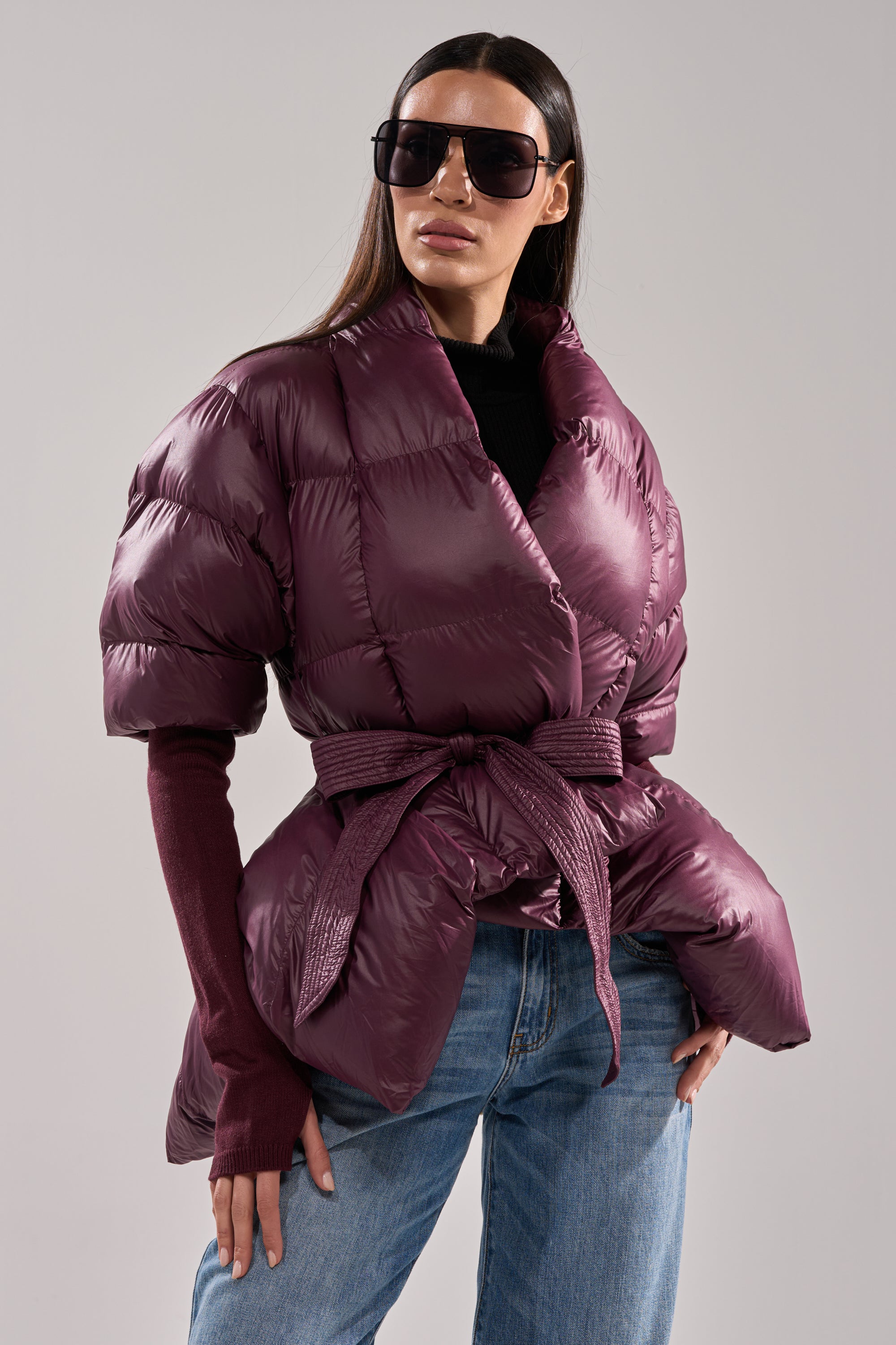 A woman models the RIB SLEEVE PEPLUM PUFFER IN PURPLE, a shiny, belted jacket, styled over a dark long-sleeve top and blue jeans with oversized dark sunglasses against a plain background. 0