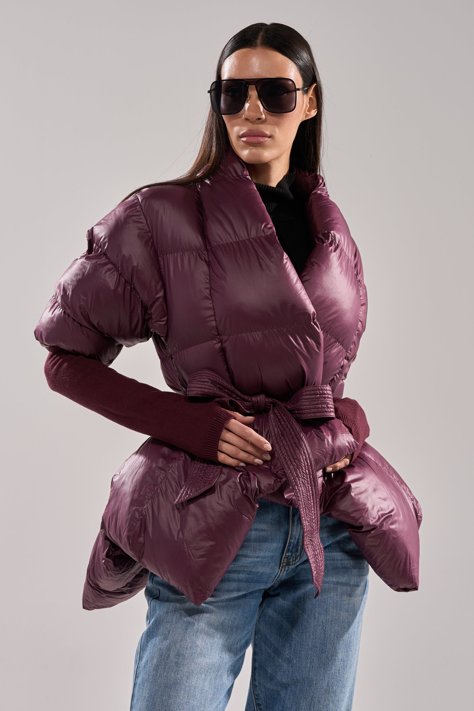 A woman confidently poses against a gray background wearing the RIB SLEEVE PEPLUM PUFFER IN PURPLE with a belt, oversized sunglasses, fingerless gloves, and blue jeans. 1