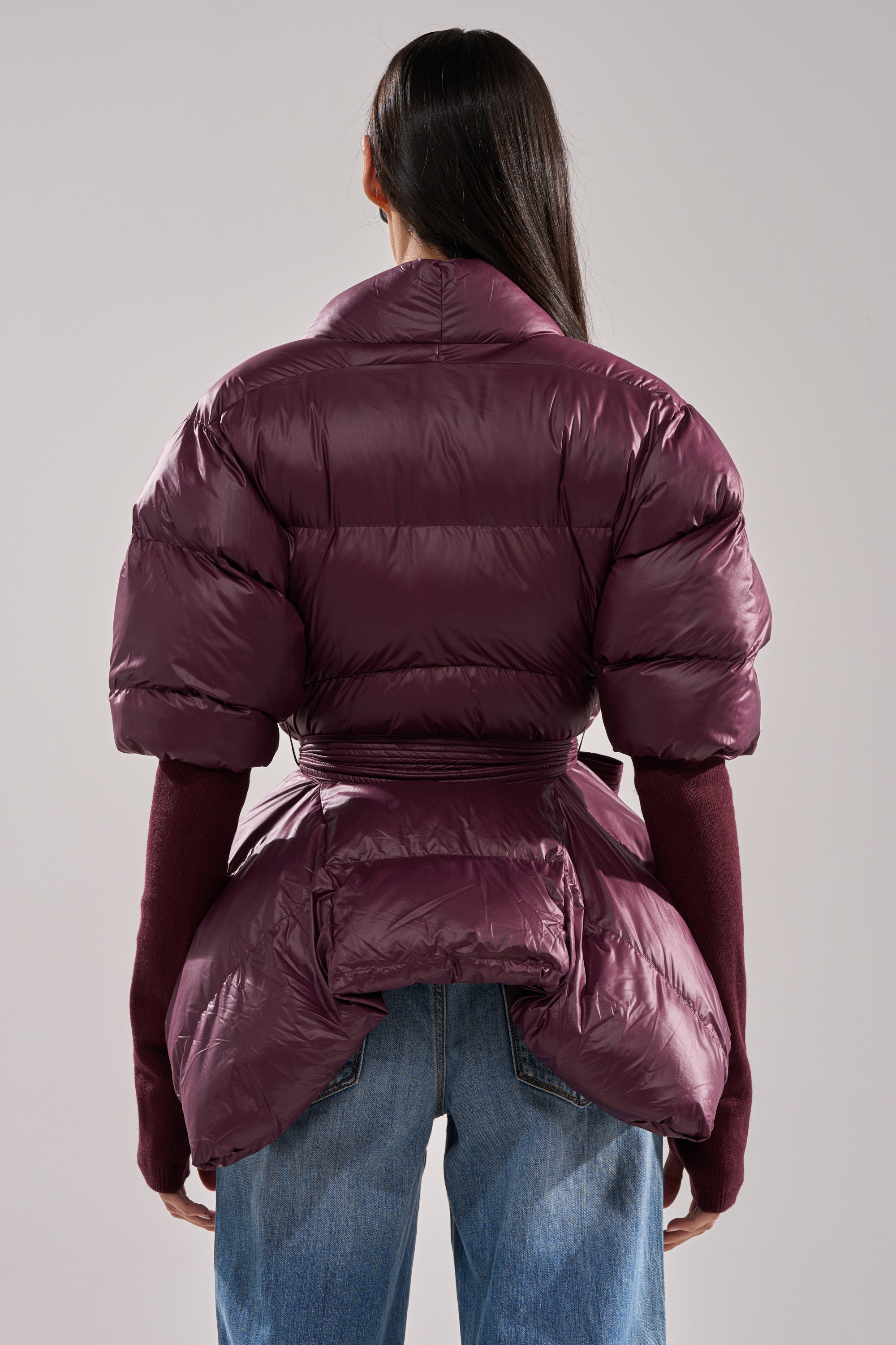 A person with long dark hair is shown from the back against a plain background, wearing the RIB SLEEVE PEPLUM PUFFER IN PURPLE over a burgundy long-sleeve top and blue jeans. 3