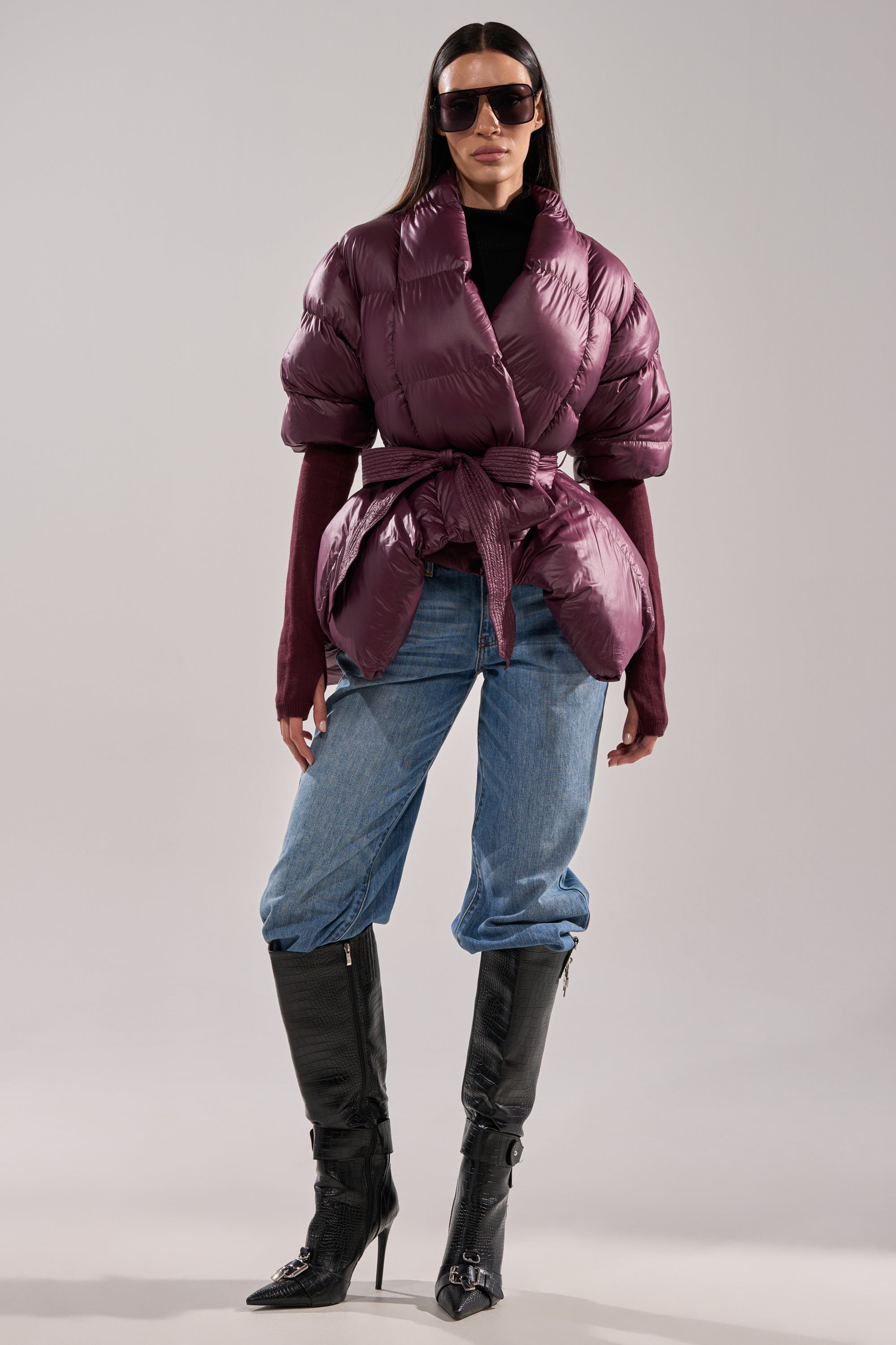 A woman in the RIB SLEEVE PEPLUM PUFFER IN PURPLE, blue cuffed jeans, black stiletto boots, a black turtleneck, and sunglasses poses against a plain gray background. 4