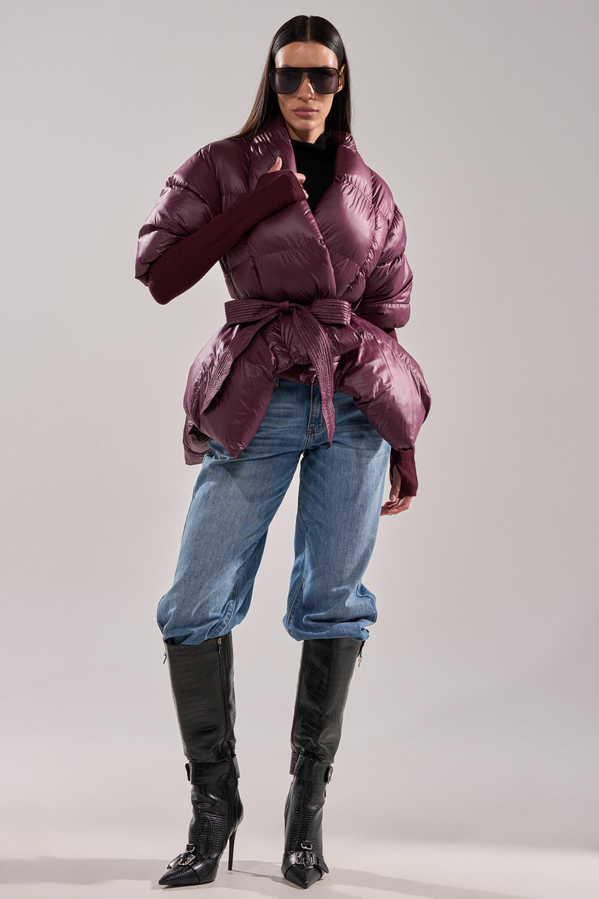 A woman stands confidently in the RIB SLEEVE PEPLUM PUFFER IN PURPLE, paired with oversized sunglasses, a black top, blue jeans tucked into black heeled boots, against a plain background. 5