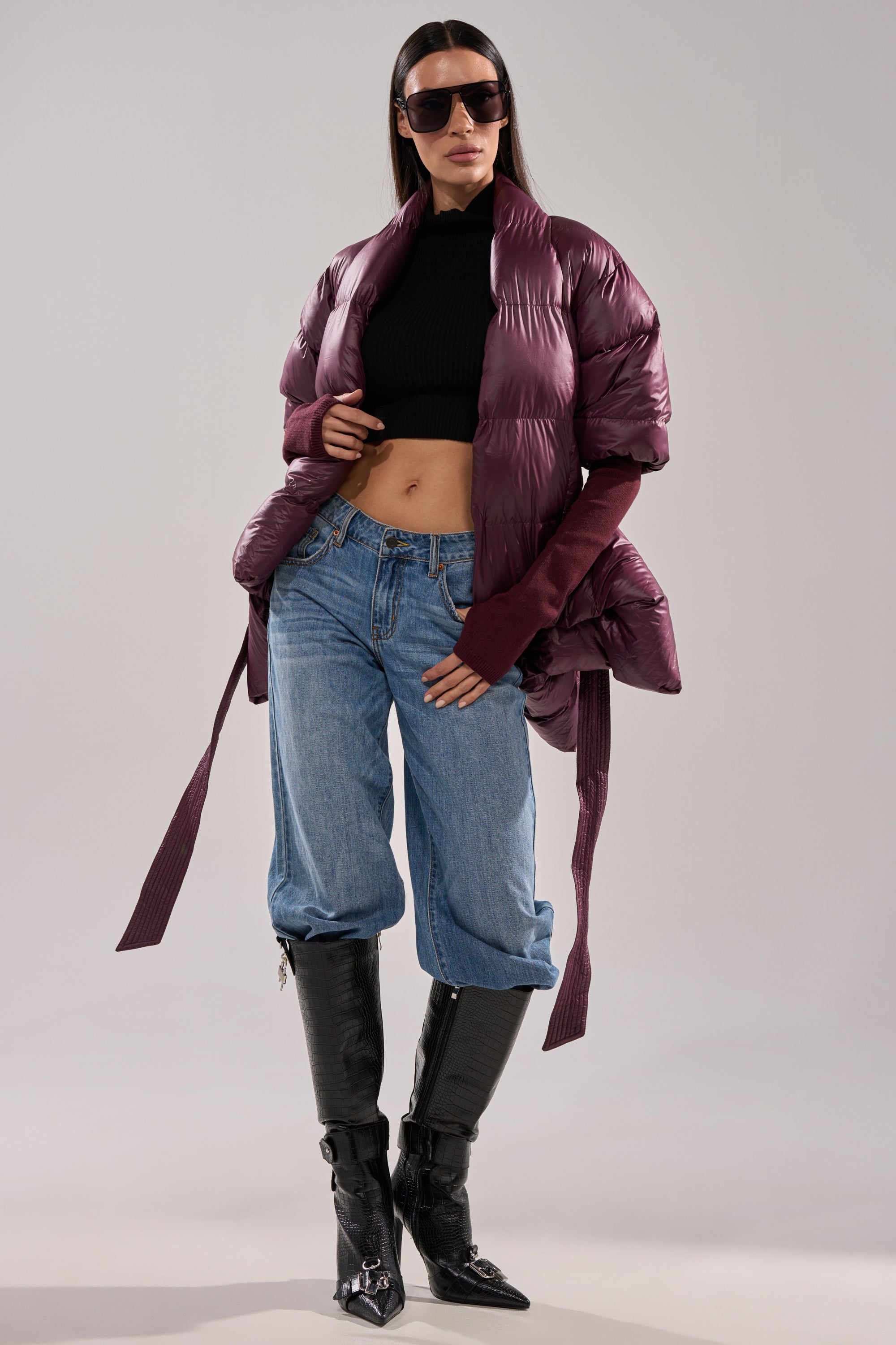 A woman wearing the RIB SLEEVE PEPLUM PUFFER IN PURPLE, a cropped black turtleneck, loose blue jeans tucked into black knee-high boots, and large sunglasses poses confidently against a plain background. 6