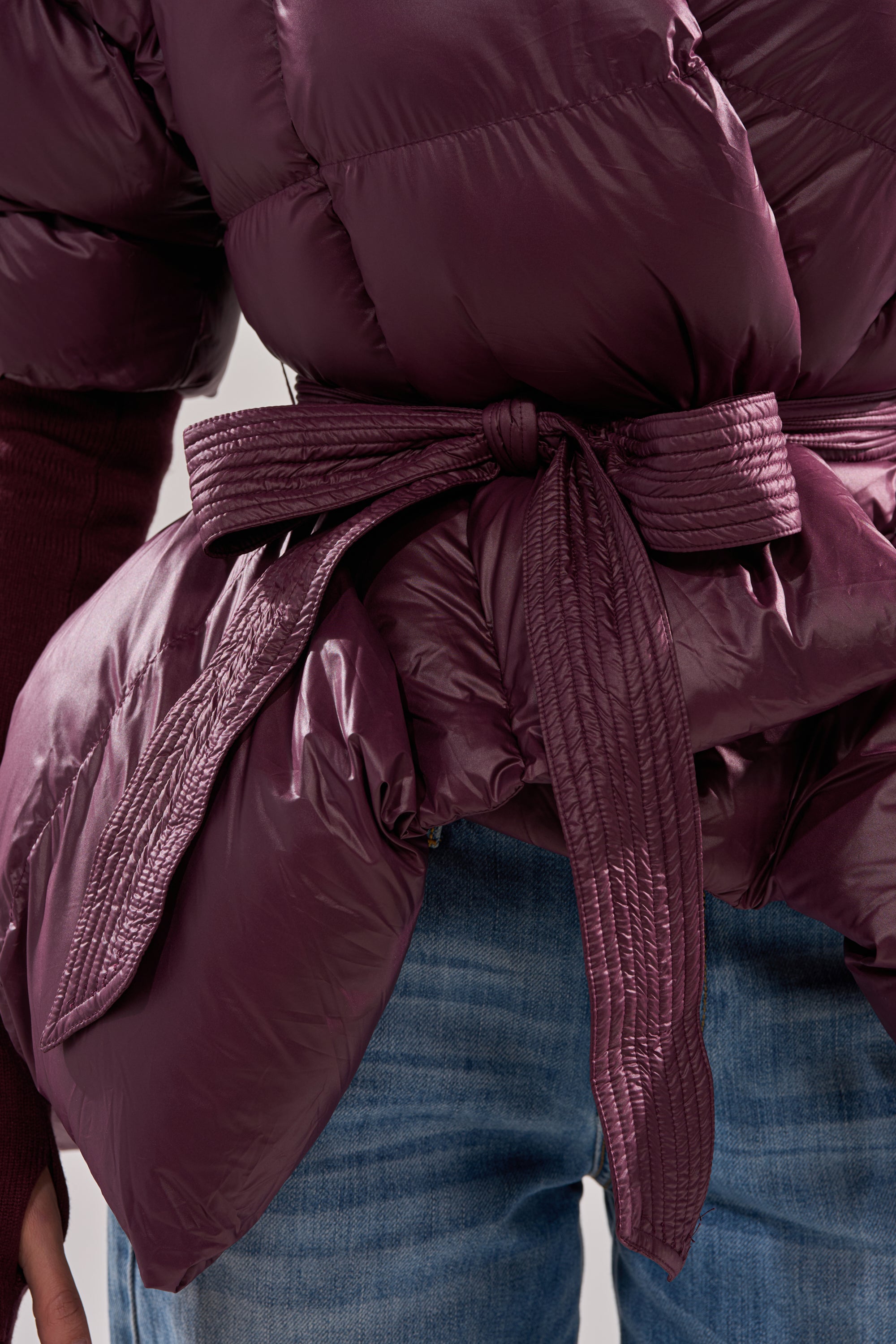 Close-up of a person wearing the RIB SLEEVE PEPLUM PUFFER IN PURPLE, a shiny dark purple jacket with a peplum silhouette and matching waist belt, styled with blue jeans and a long-sleeve top for a chic winter look. 7