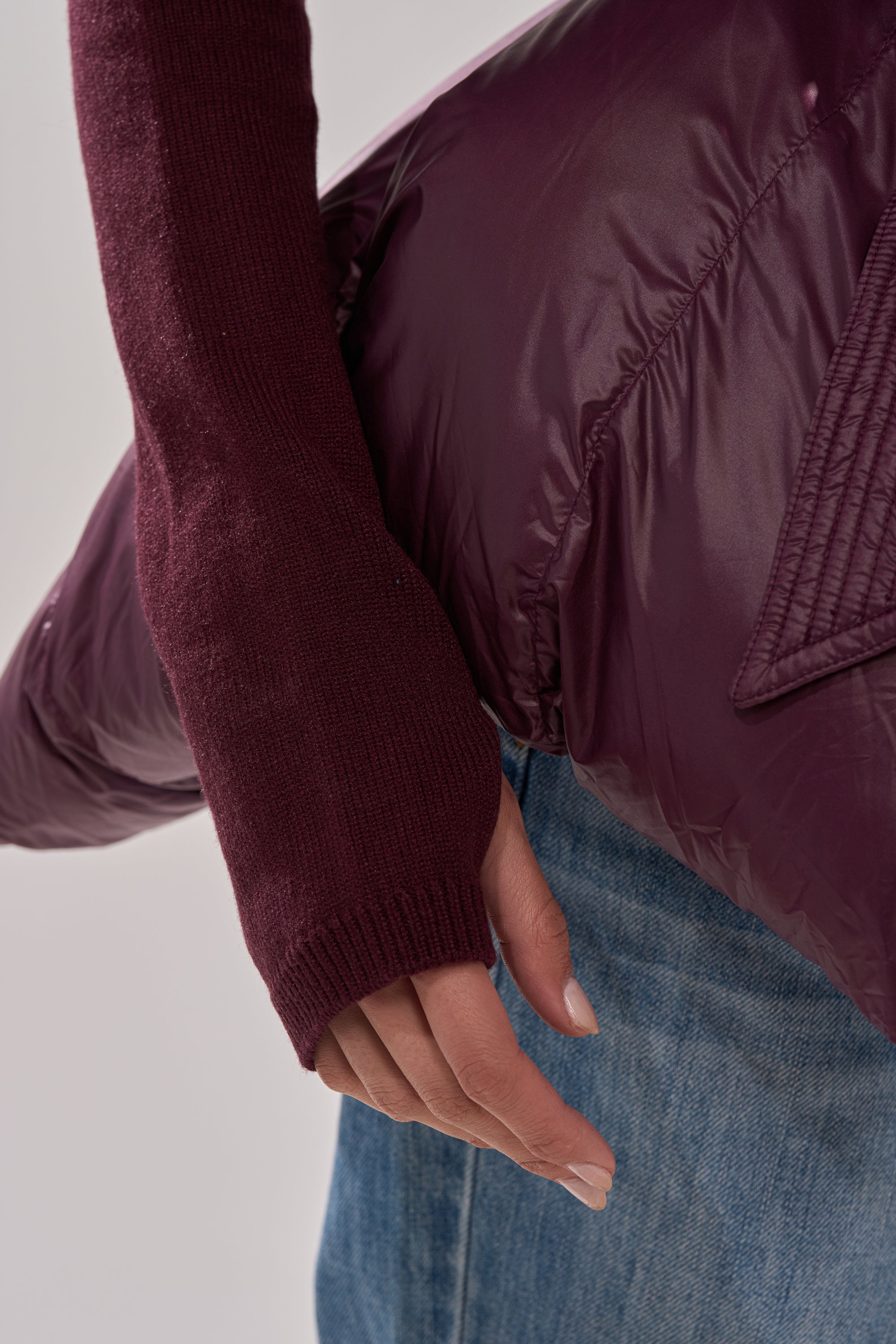 A person in a maroon sweater and blue jeans holds the RIB SLEEVE PEPLUM PUFFER IN PURPLE over their arm, with focus on their hand and part of the jacket. 8