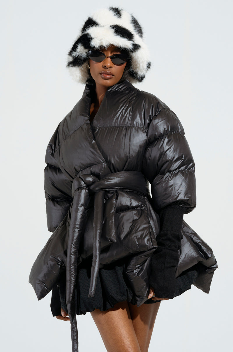 A person wearing a shiny, oversized black A Step Ahead Long Ribbed Sleeve Peplum Puffer Coat with a cocoon collar and belt, along with black gloves and a black-and-white fur hat. They are also sporting sunglasses and a black skirt, standing against a plain light-colored background.