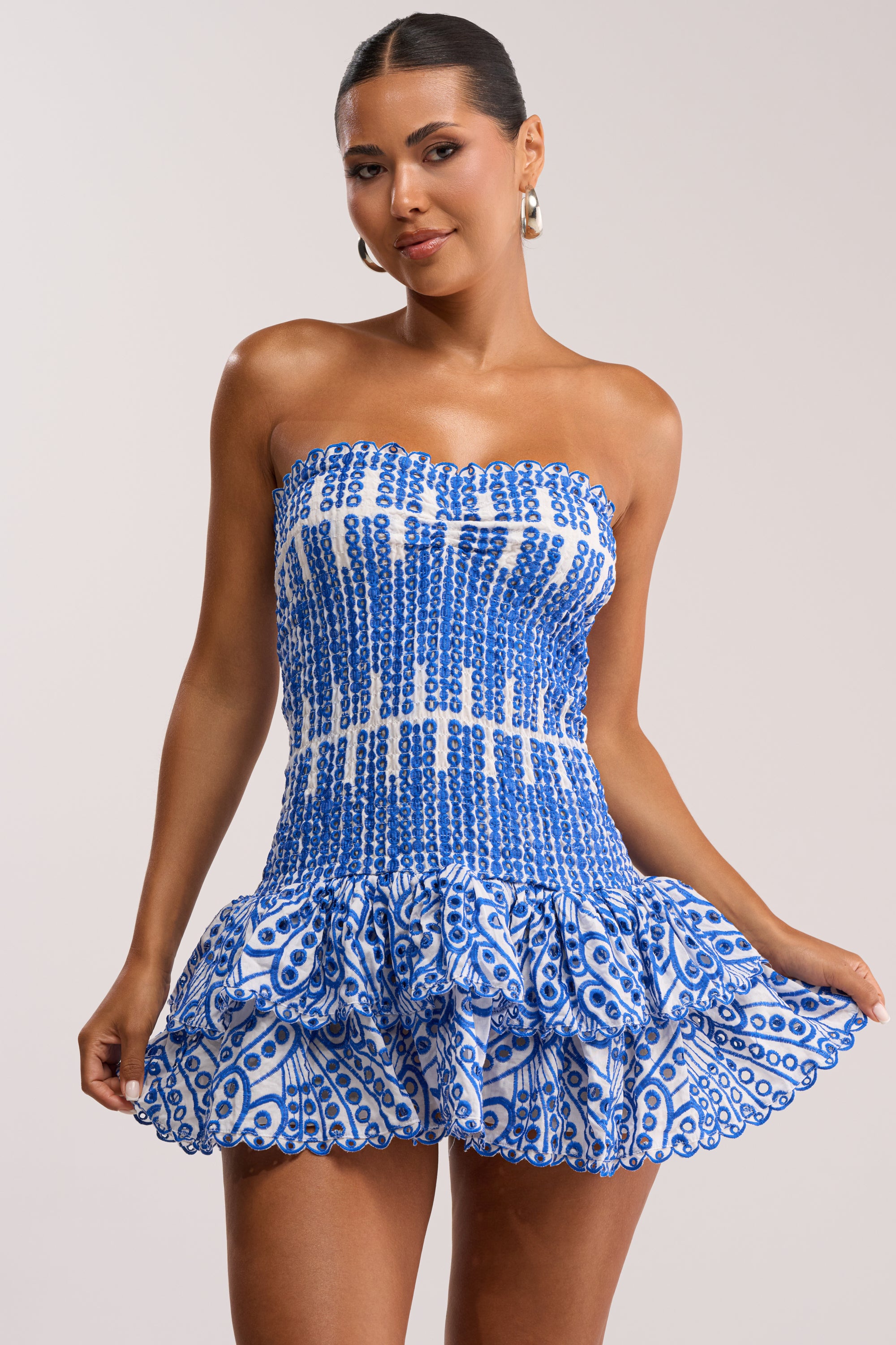 A woman wears the SUMMER OF LOVE SMOCKED EYELET STRAPLESS MINI DRESS, featuring a blue and white geometric pattern and ruffled skirt, perfect for summer. She styles it with slicked back hair and large hoop earrings.