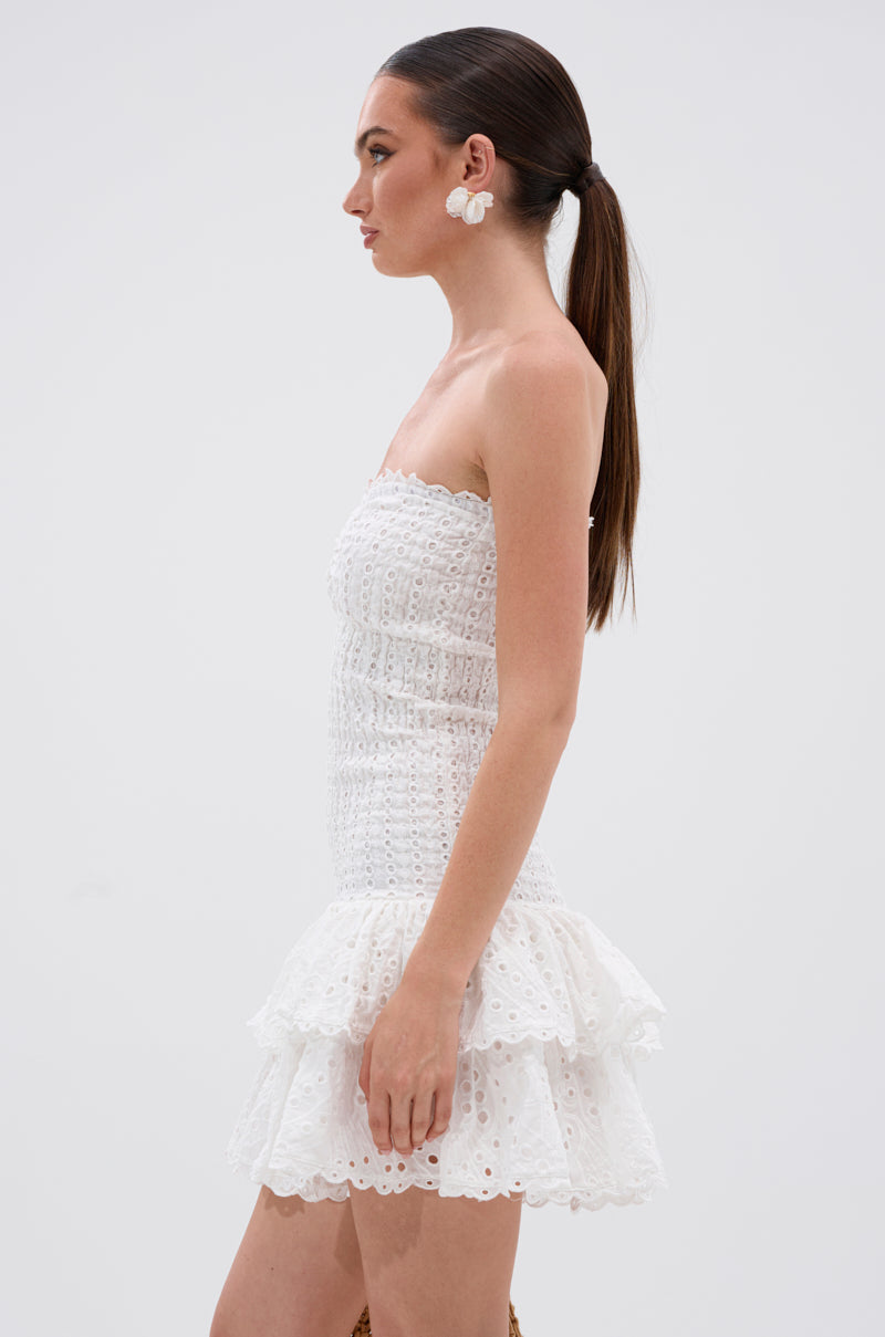 Woman wears the LETS GO TO THE BEACH RUFFLE MINI DRESS, a strapless white eyelet lace mini with ruffles and a skater silhouette, styled with large white floral earrings and a low ponytail, posing in profile against a plain background.