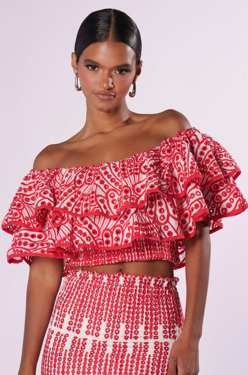 A woman stands against a plain background, wearing the SUMMER LOVING OFF THE SHOULDER RUFFLE CROP BLOUSE paired with a matching skirt. The outfit showcases intricate patterns, and she has her hair pulled back, accessorized with hoop earrings. Perfect as a summer top, this ensemble exudes effortless style and charm.