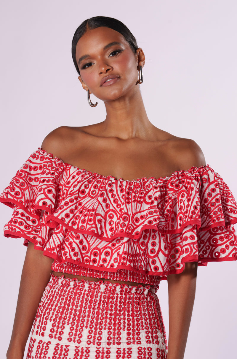 A woman with dark hair is wearing hoop earrings and the SUMMER LOVING OFF THE SHOULDER RUFFLE CROP BLOUSE paired with a matching skirt. The red and white ensemble showcases intricate patterns featuring circular and elongated designs. Standing confidently against a light neutral background, she epitomizes summer elegance.