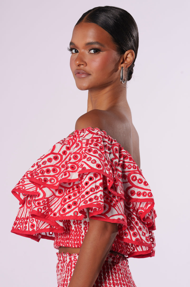 A person with neatly pulled-back hair is wearing the SUMMER LOVING OFF THE SHOULDER RUFFLE CROP BLOUSE, a colorful off-the-shoulder top featuring multiple layers of ruffles in red and white patterns. They are posing with a confident expression against a plain background.