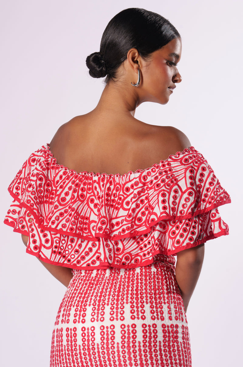A person stands with their back facing the camera, showcasing the SUMMER LOVING OFF THE SHOULDER RUFFLE CROP BLOUSE and a matching skirt adorned with a red and white circular pattern. Their hair is styled in a sleek bun, and they are wearing hoop earrings. The background is plain white.