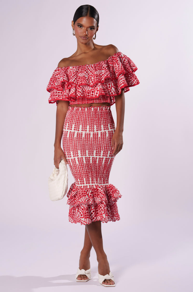 A woman stands against a light background, wearing the vibrant red and white Summer Loving Off-the-Shoulder Ruffle Crop Blouse with ruffled sleeves and eyelet patterning, paired with a matching high-waisted, knee-length skirt adorned with ruffles at the hem. She holds a small white handbag and wears white heeled sandals.