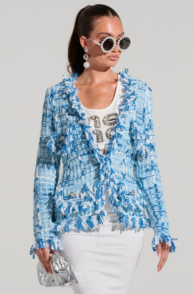 A woman wears oversized sunglasses, pearl earrings, and a white shirt with text under the SASHA BUTTON DOWN CARDIGAN SWEATER, featuring elegant fringe details. She holds a silver clutch and dons a white skirt. Her hair is tied back, exuding a fashionable and confident look.