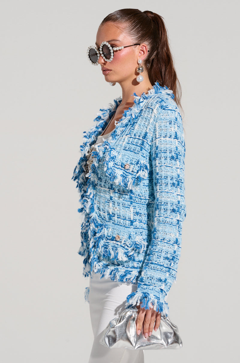 A woman with long brown hair in a high ponytail wears large, round sunglasses with decorative frames. She is dressed in the SASHA BUTTON DOWN CARDIGAN SWEATER, featuring a blue and white tweed-inspired design with fringe details, paired with white pants and holding a shiny silver clutch bag. She has dangling earrings.