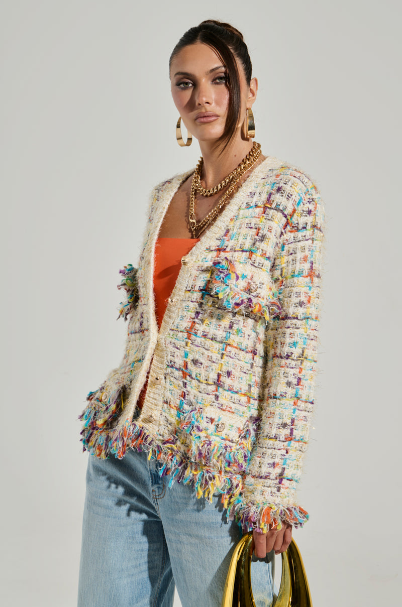 A woman stands confidently, wearing the Margaret Button Down Cardigan in Cream, a tweed-inspired piece with rainbow accents and fringed edges, over an orange top and light blue jeans. She holds a gold handbag and has her hair styled in a sleek partial updo. Completing her look, she accessorizes with large hoop earrings and layered gold necklaces.