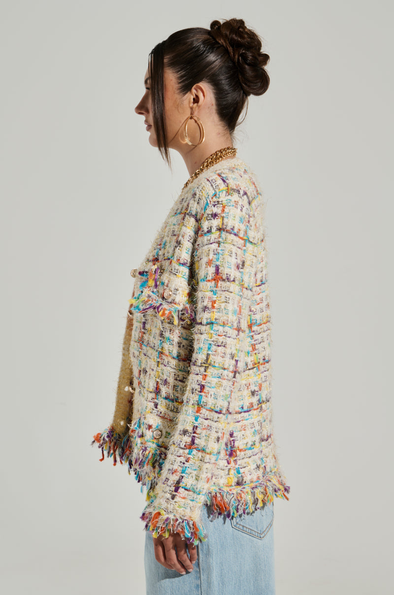 A woman with her hair in a bun is shown from the side, wearing the MARGARET BUTTON DOWN CARDIGAN IN CREAM, which features colorful woven patterns and fringe details at the hem, cuffs, and front pockets. The cardigan is embellished with metallic gold buttons. She is also wearing large hoop earrings and light blue jeans against a plain, neutral background.