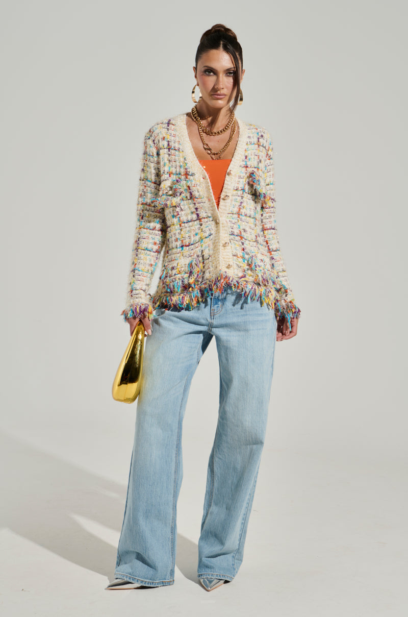 A woman poses against a plain backdrop, wearing the MARGARET BUTTON DOWN CARDIGAN IN CREAM with fringe detailing over an orange top, light blue wide-leg jeans, and silver pointed-toe heels. She holds a gold handbag and accessorizes with gold earrings and layered necklaces.