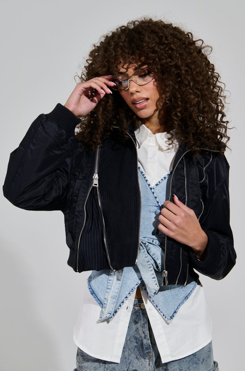 A person with curly hair wearing glasses and the modern DUKE ZIPPER DETAIL BOMBER over a white shirt and denim overalls stands against a plain background. They hold the edge of their glasses with one hand, gazing down thoughtfully.