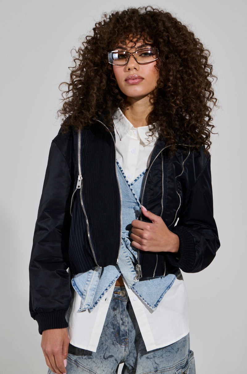 With curly hair and fashionable glasses, a person confidently sports the DUKE Zipper Detail Bomber over a white shirt, layered under chic denim overalls. Against a plain backdrop, they effortlessly embody trendiness.