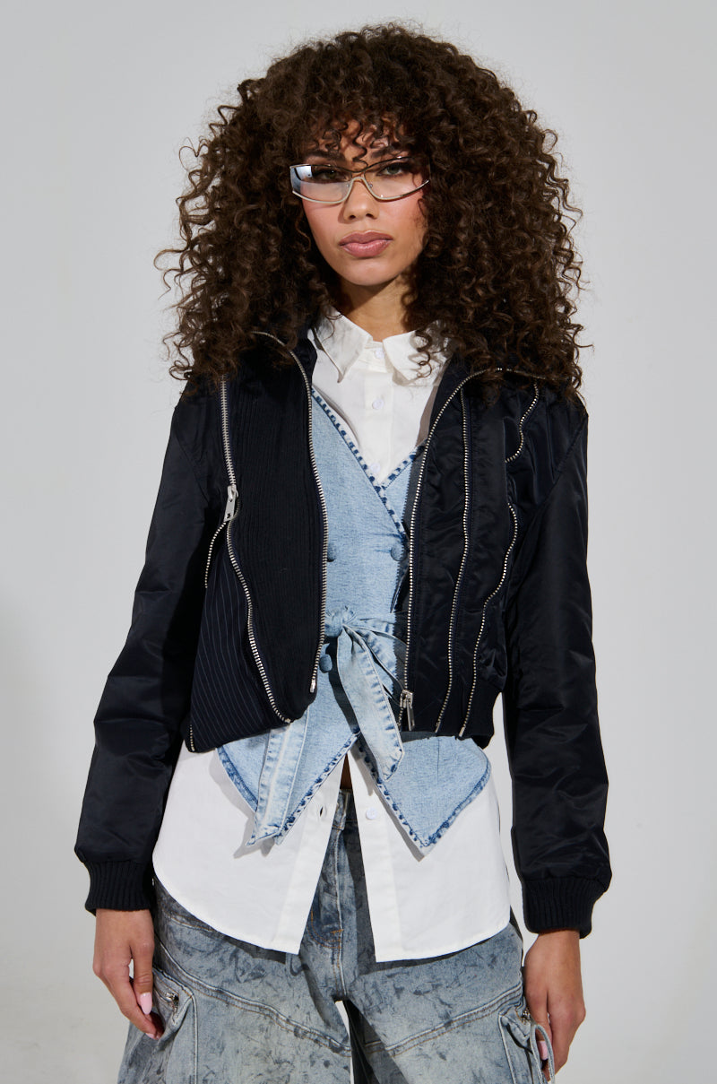 A person with curly hair is wearing the DUKE ZIPPER DETAIL BOMBER jacket over a layered white shirt and denim vest. They have on light-wash denim pants with patchwork details. The individual, who is also wearing glasses, is facing the camera in a neutral pose against a plain background.