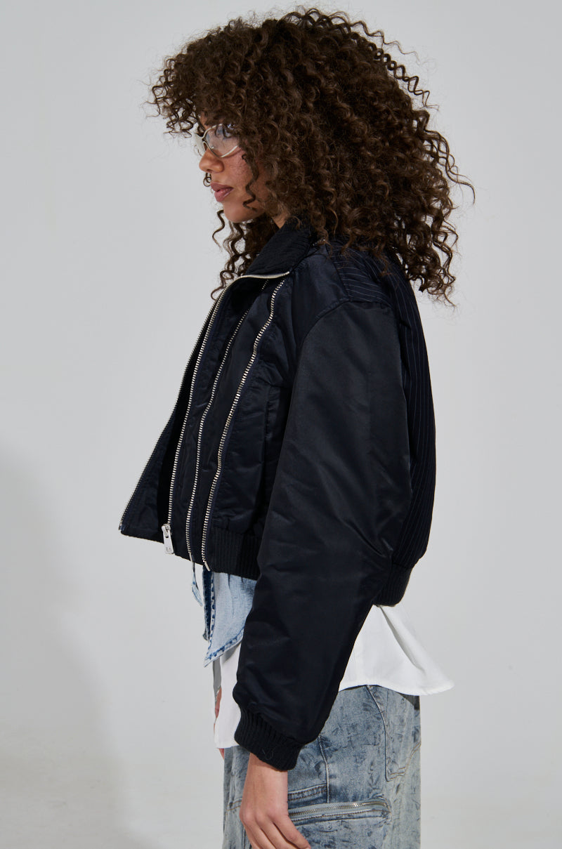 A person with curly hair is wearing the DUKE ZIPPER DETAIL BOMBER, a black cropped jacket with zipper accents, over a white shirt. They are paired with denim pants that feature a light pattern and are accessorizing with clear glasses. The person is standing sideways, facing left against a plain background.