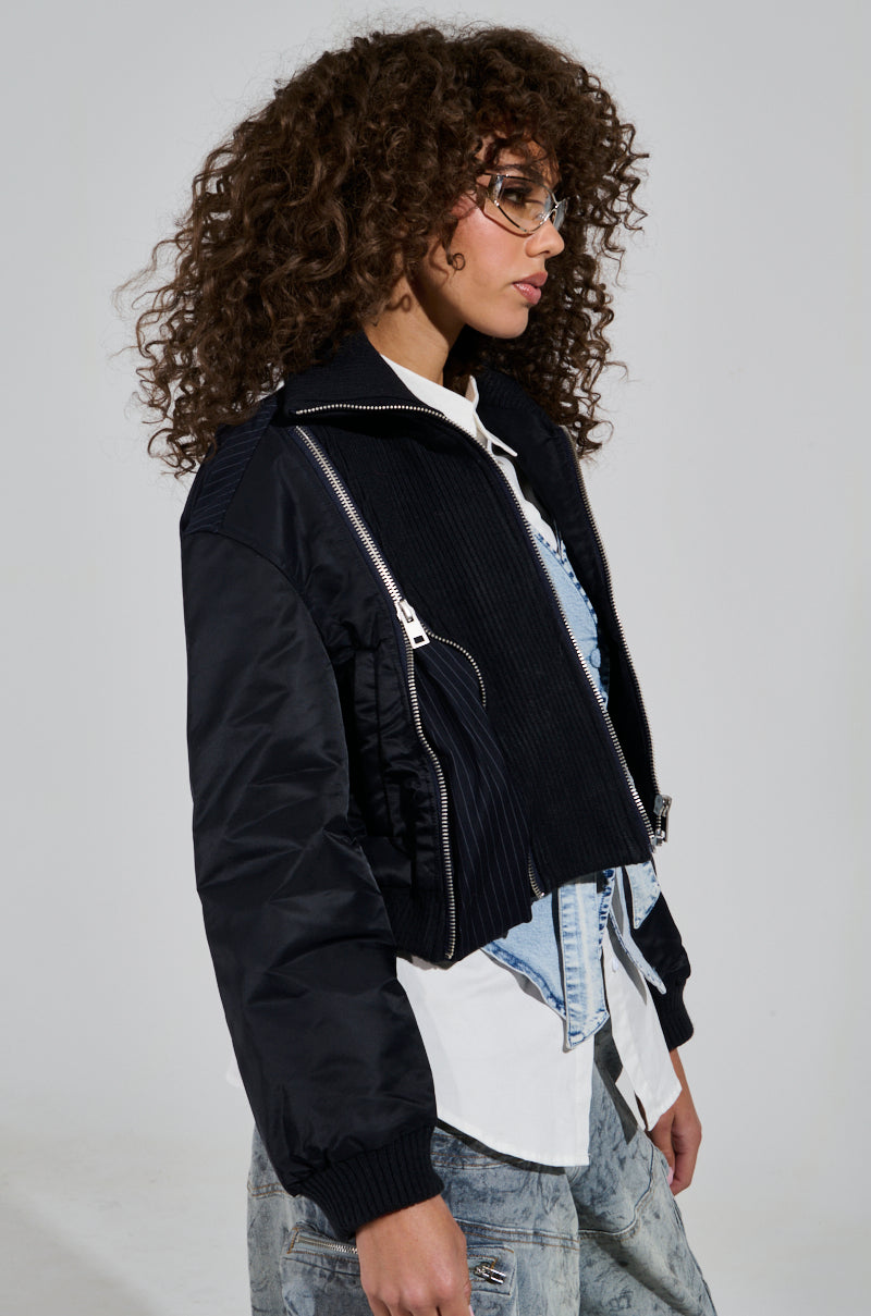 A person with curly hair is wearing the trendy DUKE ZIPPER DETAIL BOMBER jacket in black over a denim vest and white shirt. They are also sporting clear glasses while standing against a plain background.