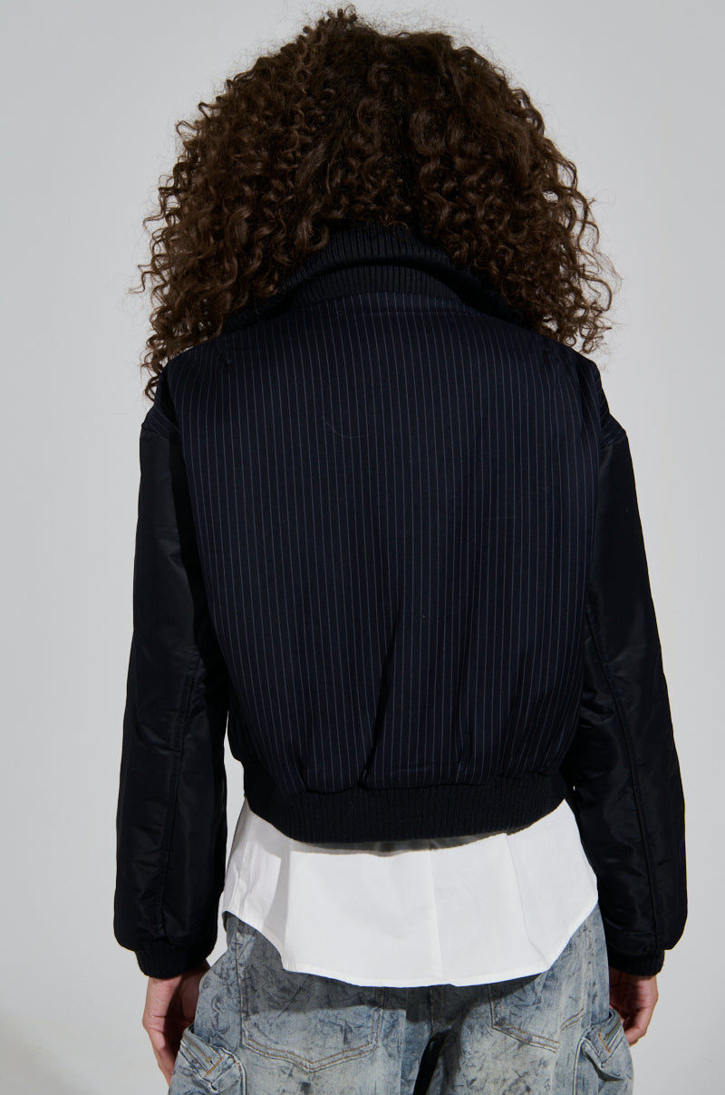 From behind, a person with curly hair wears the DUKE ZIPPER DETAIL BOMBER, a dark pinstriped jacket featuring zipper details, over a white shirt with a frilled hem. They also have on faded blue jeans against a plain, light-colored background.
