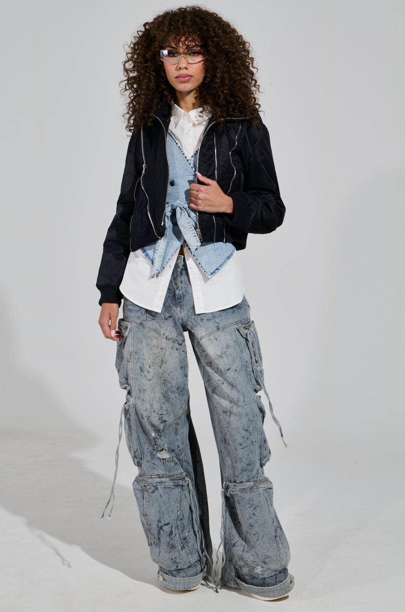 A person with curly hair and glasses is dressed in the DUKE ZIPPER DETAIL BOMBER, featuring a black color. They have layered shirts and a vest, complemented by oversized distressed cargo pants and sneakers, standing against a plain background.
