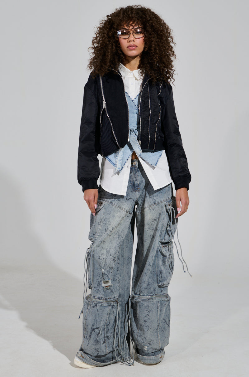 A person with curly hair and glasses stands in a fashion-forward outfit, featuring the DUKE Zipper Detail Bomber layered over a white shirt and a blue vest. Paired with baggy, textured jeans, their look is effortlessly casual and edgy.