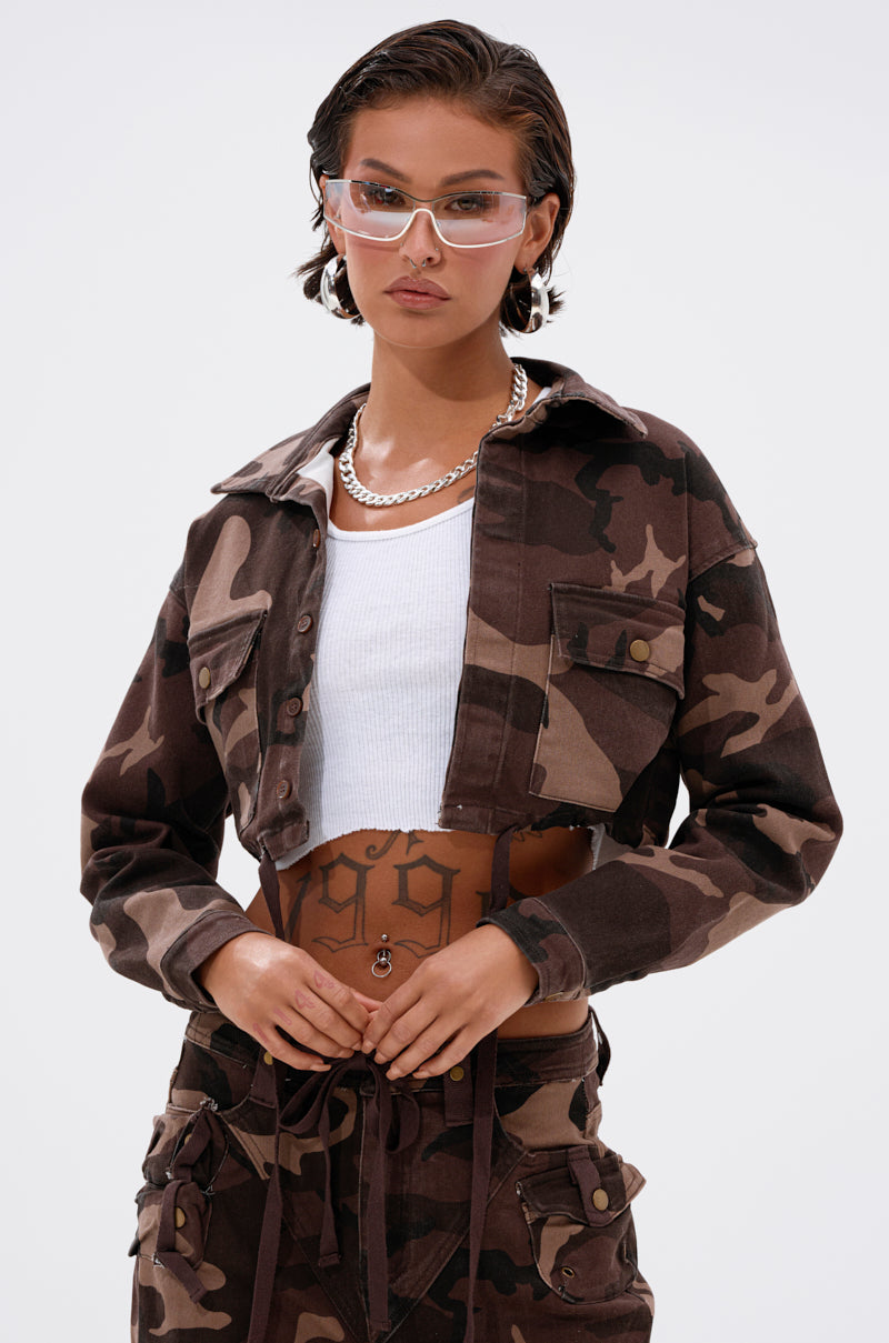 A person wears the CANT SEE ME BROWN CAMO CROPPED BUTTON DOWN with matching pants, a white ribbed crop top, clear sunglasses, a silver chain necklace, and shows off a tattooed midriff featuring "1999" on the stomach. 0