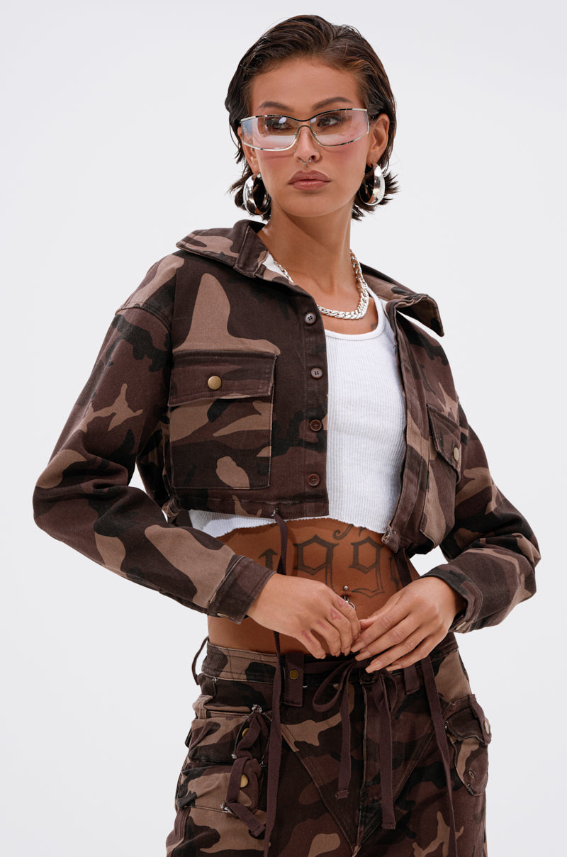 Wearing the CANT SEE ME BROWN CAMO CROPPED BUTTON DOWN with matching camo pants, clear sunglasses, a pearl necklace, and a white crop top, a person poses confidently against a plain light background. 1