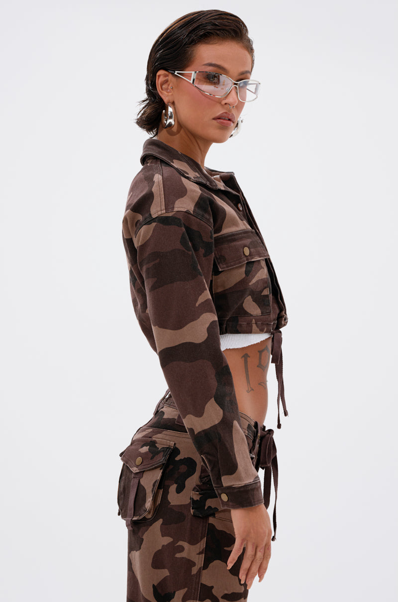 A woman with slicked-back hair and clear glasses wears the CANT SEE ME BROWN CAMO CROPPED BUTTON DOWN from AKIRA Label, paired with matching cargo pants. She accessorizes with large hoop earrings and displays a torso tattoo, posed against a white background. 3