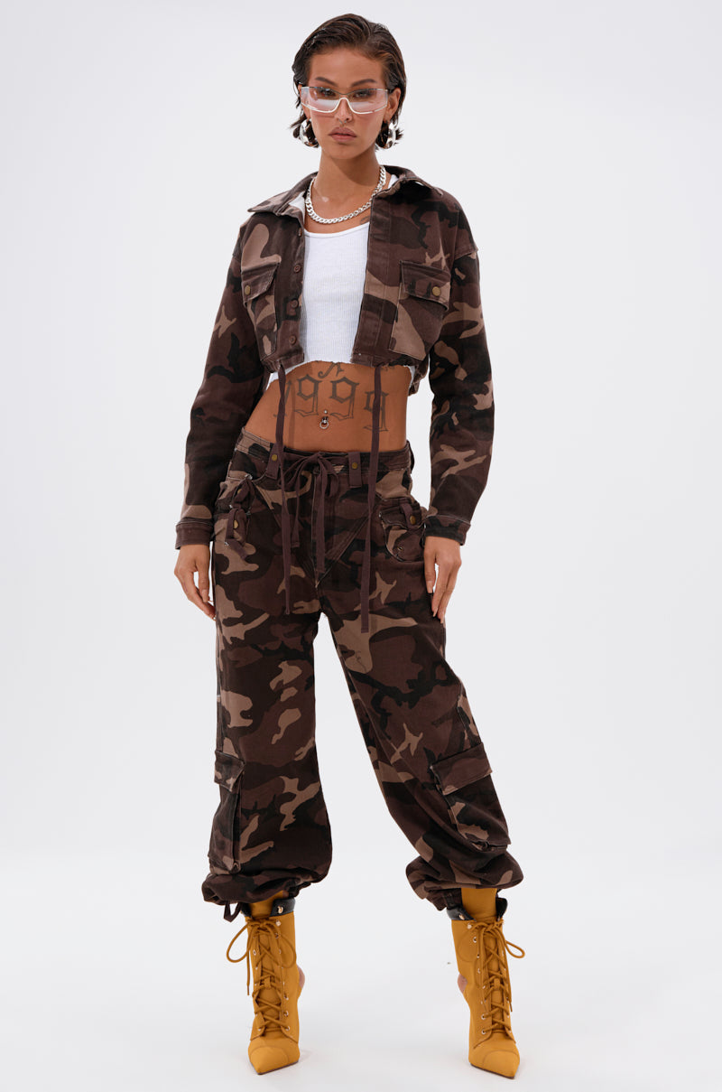 Wearing the CANT SEE ME BROWN CAMO CROPPED BUTTON DOWN with matching pants, a white crop top, yellow lace-up boots, clear-framed glasses, and layered necklaces, a person stands confidently against a plain white background. 7