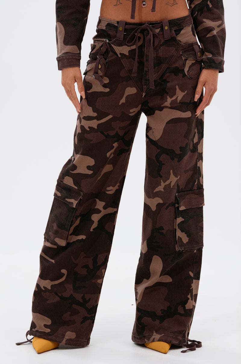 A person wears the CANT SEE ME BROWN CAMP CARGO JOGGER—wide-leg camo pants with a drawstring waist and large side pockets—paired with yellow pointed-toe shoes. Only the lower torso and legs are shown.