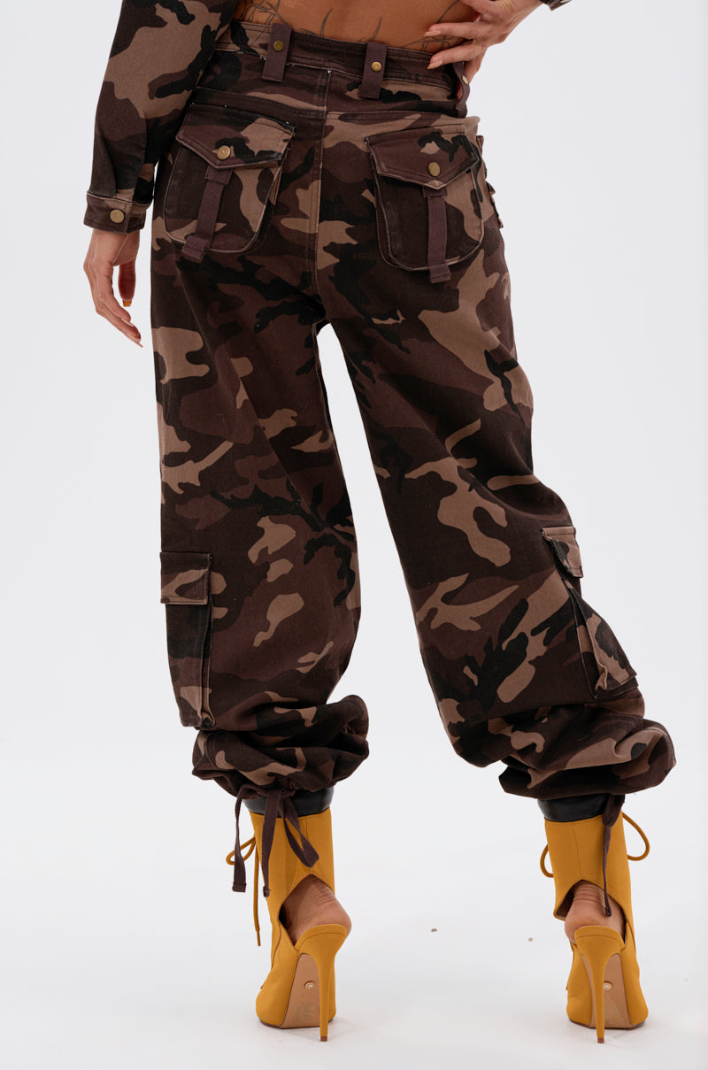 A person stands facing away, wearing the CANT SEE ME BROWN CAMP CARGO JOGGER with multiple pockets, a matching cropped jacket, and bright yellow high-heeled boots against a plain white background.
