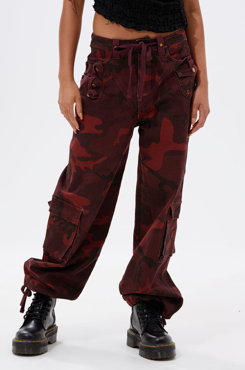 A person stands wearing the CANT SEE ME BURGUNDY CAMP CARGO JOGGER, featuring loose-fitting burgundy cargo style with multiple pockets and buttons, paired with black lace-up combat boots. Upper body is not visible. 2