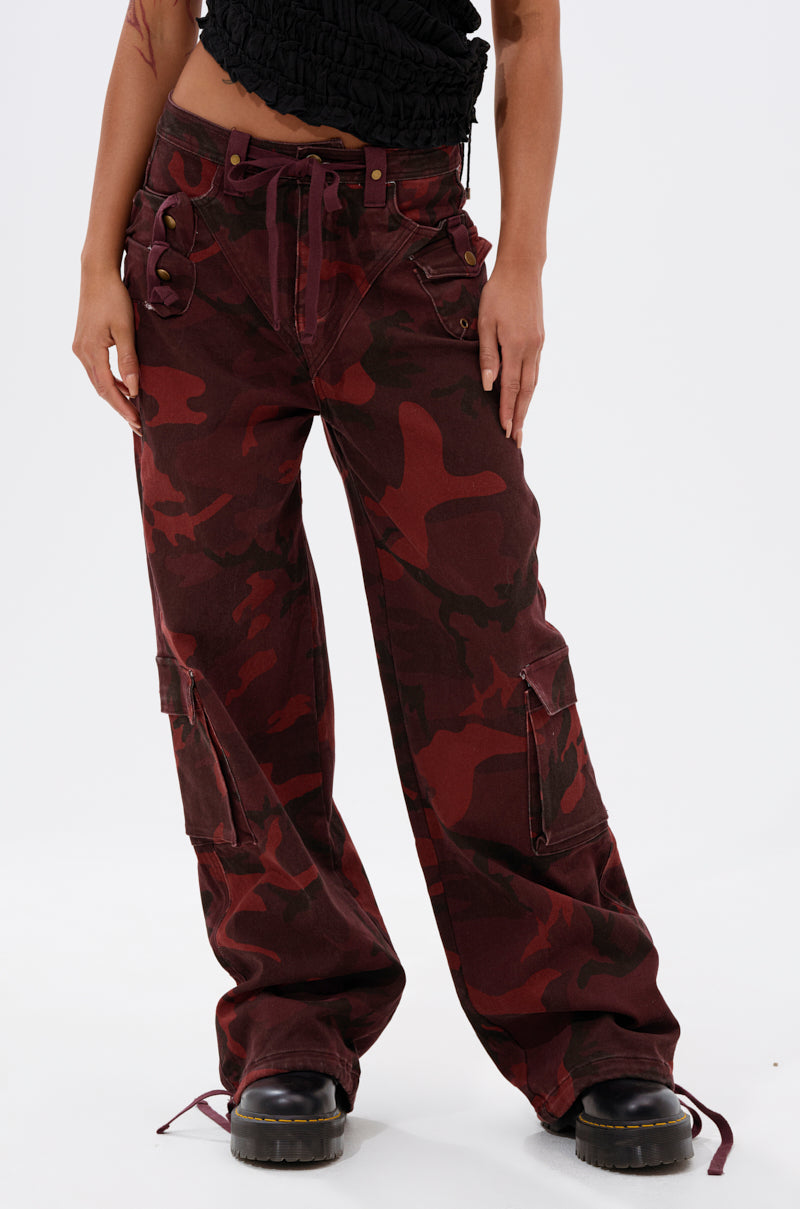 Model wears the CANT SEE ME BURGUNDY CAMP CARGO JOGGER—high-waisted, loose cargo joggers with a red and dark camo print and multiple pockets—styled with black chunky boots against a white background. 3