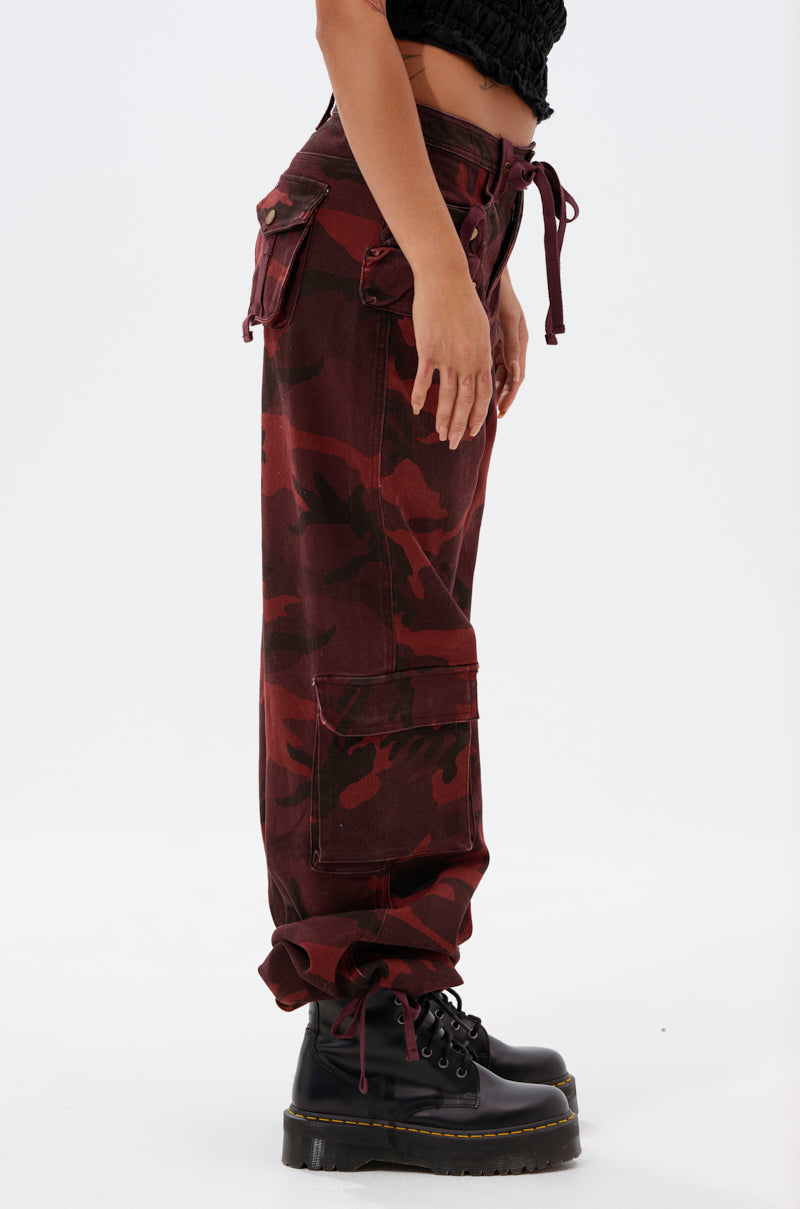 A person stands sideways in the CANT SEE ME BURGUNDY CAMP CARGO JOGGER with side pockets, paired with black platform combat boots and a black top, creating a bold streetwear vibe against a plain white background. 4