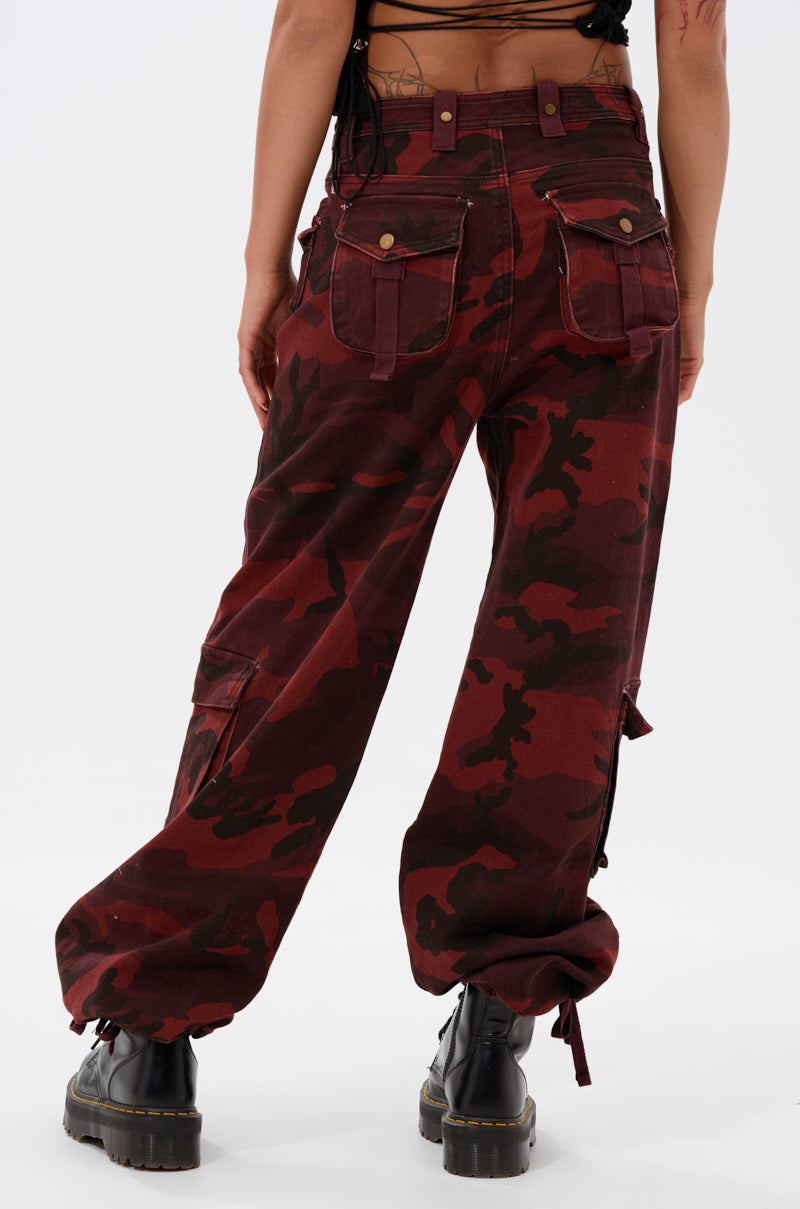 Wearing the CANT SEE ME BURGUNDY CAMP CARGO JOGGER with multiple pockets, this person stands with their back to the camera. The baggy joggers are styled with black lace-up boots for an edgy urban vibe. 5