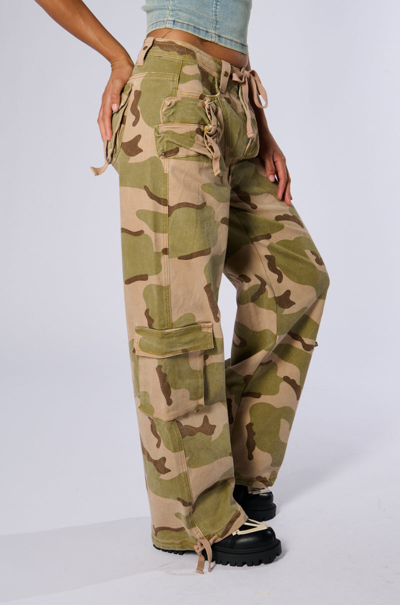 Side view of a person wearing the ON TOP OF IT CAMO JOGGER, featuring high-rise design, multiple flap pockets, and lace-up detailing. They are also sporting a cropped top and black chunky-heeled boots against a plain white background.